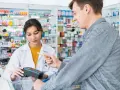 Klient płaci kartą w aptece, gdzie farmaceutka obsługuje terminal. W tle widać półki z lekami, a na nich produkt z napisem ursopol.
