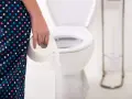 Osoba w kropkowanej sukience trzymająca rolkę papieru toaletowego przed toaletą.