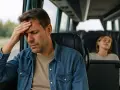 Mężczyzna z ręką na czole w autobusie, cierpiący na chorobę lokomocyjną. Obok śpi młoda kobieta.