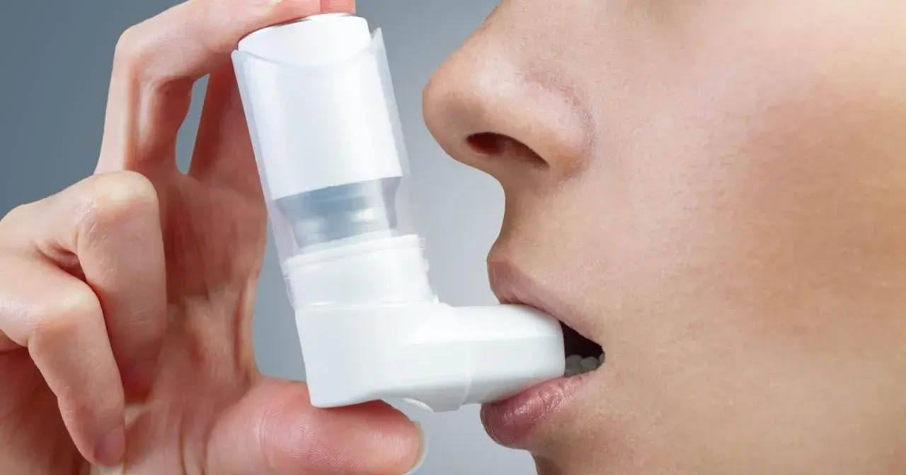 Dłoń trzyma biały inhalator, zbliżając go do ust osoby. To pomoc w oddychaniu, jak Ventolin aerozol.