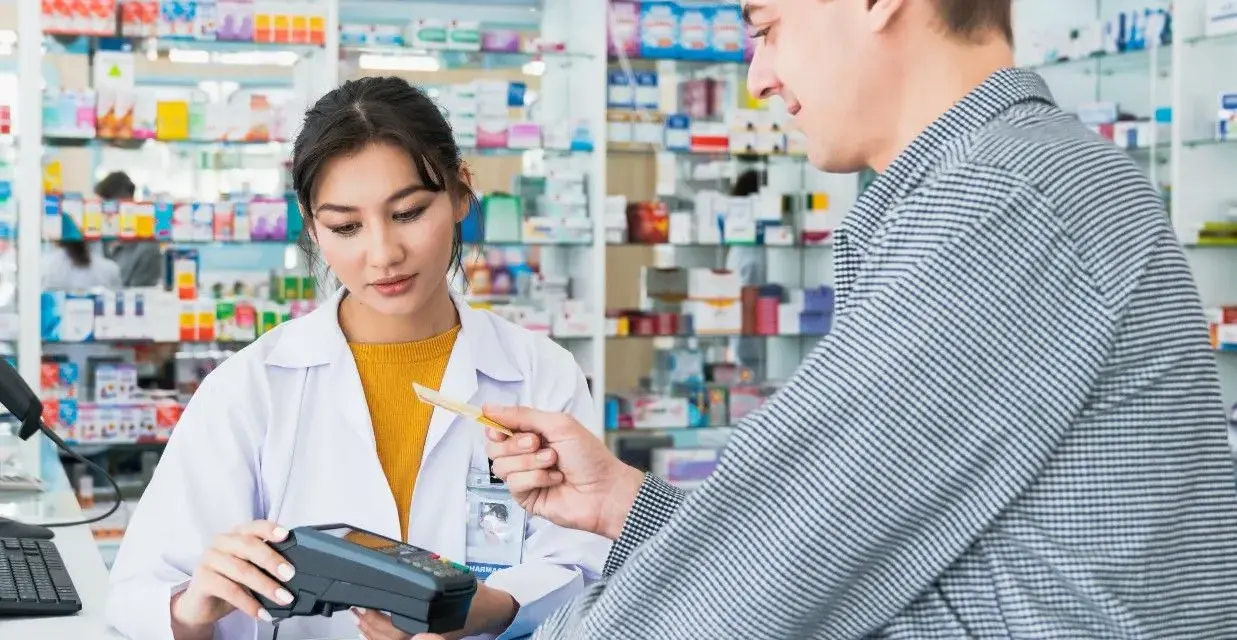Klient płaci kartą w aptece, gdzie farmaceutka obsługuje terminal. W tle widać półki z lekami, a na nich produkt z napisem ursopol.