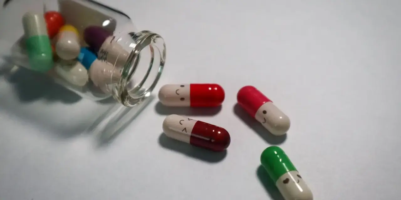 Kolorowe kapsułki, w tym leki psychotropowe, wysypują się ze szklanej buteleczki. Niektóre mają narysowane uśmiechnięte buźki.