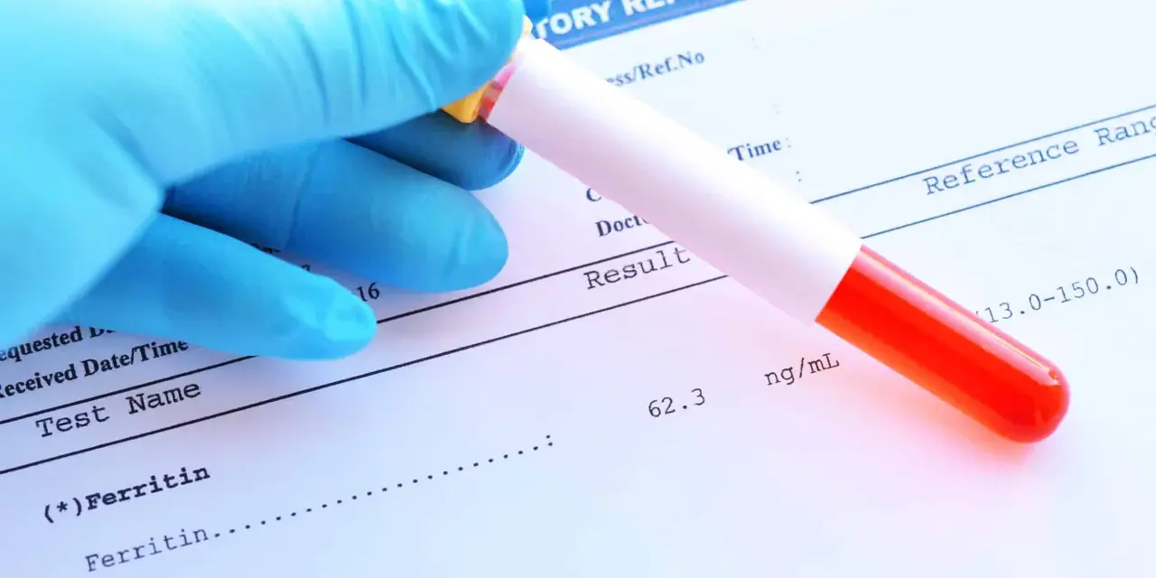 Ręka w niebieskiej rękawiczce trzyma probówkę z krwią nad raportem laboratoryjnym. Wynik ferrytyny 62.3 ng/mL mieści się w normie.