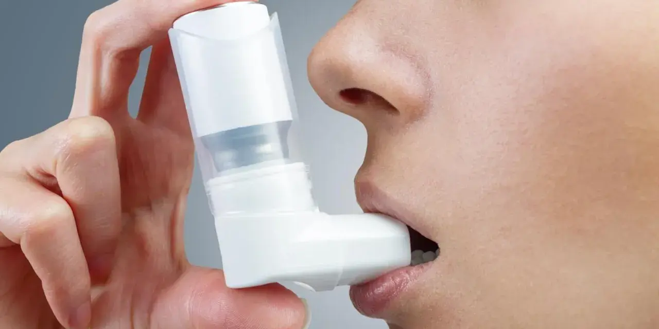 Dłoń trzyma biały inhalator, zbliżając go do ust osoby. To pomoc w oddychaniu, jak Ventolin aerozol.