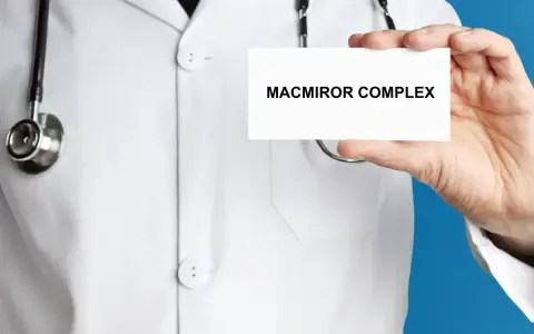 Lekarz w białym kitlu prezentuje kartkę z napisem MACMIROR COMPLEX. Stetoskop wisi na jego szyi.