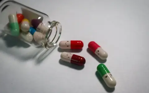 Kolorowe kapsułki, w tym leki psychotropowe, wysypują się ze szklanej buteleczki. Niektóre mają narysowane uśmiechnięte buźki.