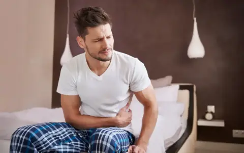 Mężczyzna siedzi na łóżku, trzymając się za brzuch. Wygląda na to, że zastanawia się, ile trwa jelitówka.