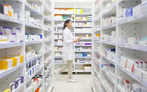 Farmaceutka w białym fartuchu idzie między regałami pełnymi leków, szukając amoksycyliny.