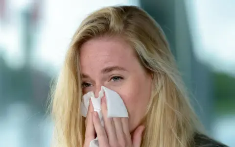 Kobieta z blond włosami chusteczką wyciera nos. Może to być objaw alergii, na którą pomoże Claritine.