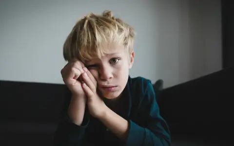 Chłopiec z blond włosami, zamyślony, z ręką przy twarzy. Jego wyraz twarzy może sugerować pewne objawy aspergera, jak trudność w nawiązywaniu kontaktu wzrokowego.