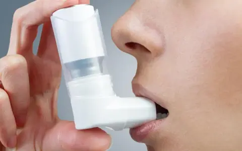 Dłoń trzyma biały inhalator, zbliżając go do ust osoby. To pomoc w oddychaniu, jak Ventolin aerozol.
