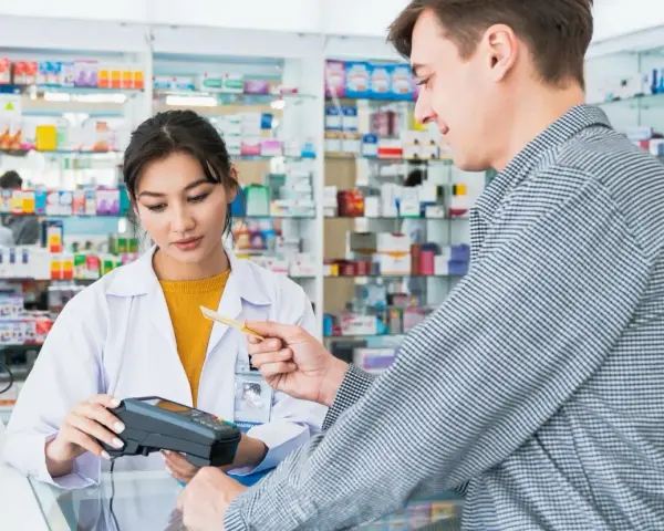 Klient płaci kartą w aptece, gdzie farmaceutka obsługuje terminal. W tle widać półki z lekami, a na nich produkt z napisem ursopol.