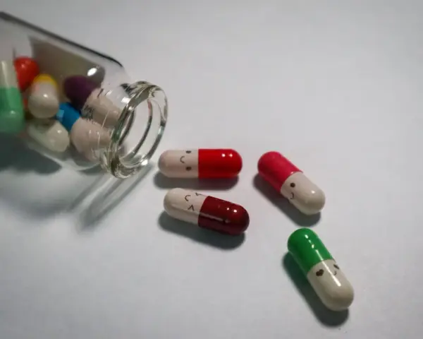 Kolorowe kapsułki, w tym leki psychotropowe, wysypują się ze szklanej buteleczki. Niektóre mają narysowane uśmiechnięte buźki.