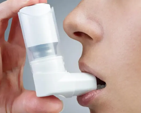 Dłoń trzyma biały inhalator, zbliżając go do ust osoby. To pomoc w oddychaniu, jak Ventolin aerozol.