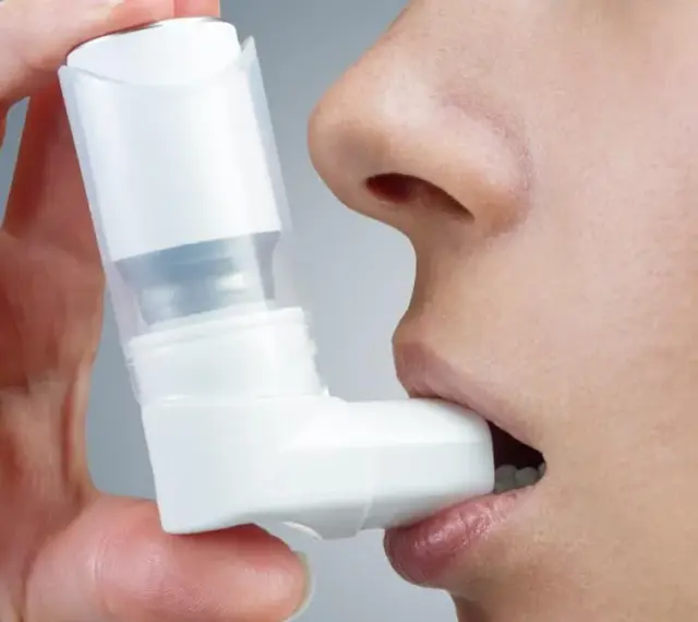 Dłoń trzyma biały inhalator, zbliżając go do ust osoby. To pomoc w oddychaniu, jak Ventolin aerozol.