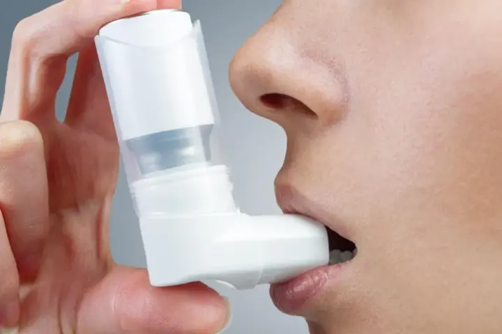 Dłoń trzyma biały inhalator, zbliżając go do ust osoby. To pomoc w oddychaniu, jak Ventolin aerozol.