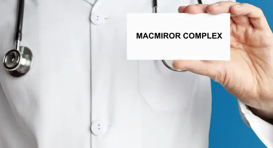 Lekarz w białym kitlu prezentuje kartkę z napisem MACMIROR COMPLEX. Stetoskop wisi na jego szyi.