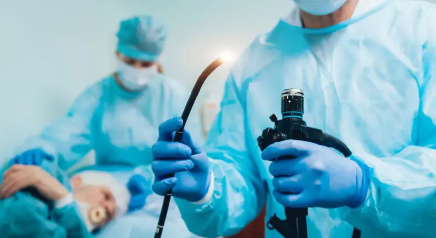 Lekarz w niebieskich rękawiczkach trzyma endoskop. W tle pacjent i inny medyk. Procedura, jak ile trwa gastroskopia, jest krótka.