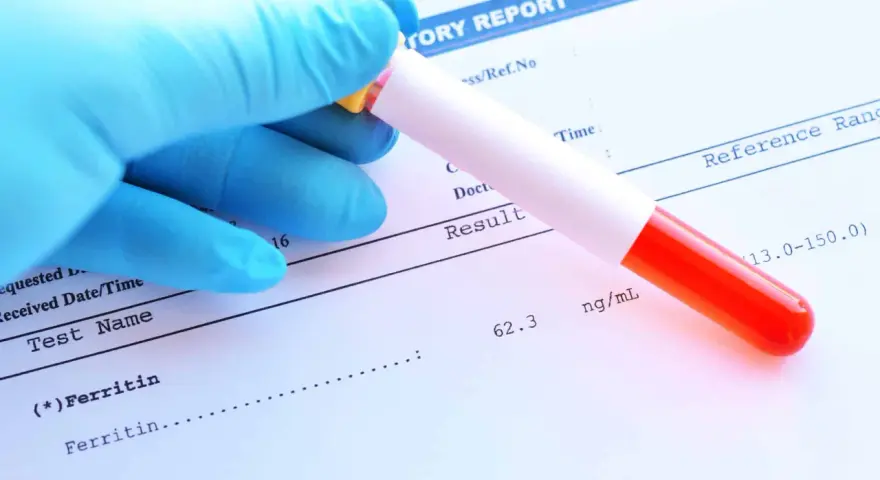 Ręka w niebieskiej rękawiczce trzyma probówkę z krwią nad raportem laboratoryjnym. Wynik ferrytyny 62.3 ng/mL mieści się w normie.