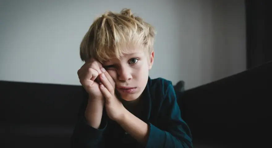 Chłopiec z blond włosami, zamyślony, z ręką przy twarzy. Jego wyraz twarzy może sugerować pewne objawy aspergera, jak trudność w nawiązywaniu kontaktu wzrokowego.